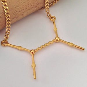 Op de afbeelding: Goudkleurige ketting met een unieke Y-vormige hanger. De hanger heeft een golvende centrale balk met drie rechte, langwerpige armen. De ketting heeft een klassiek curb link ontwerp.