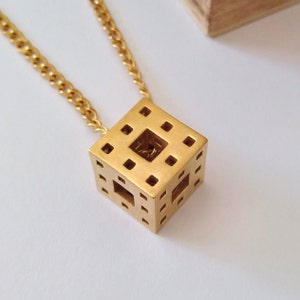 Può includere: Una collana d'oro con un ciondolo a forma di cubo geometrico. Il cubo presenta un motivo ripetitivo di quadrati ritagliati, creando un effetto tridimensionale.