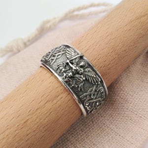 May include: A silver-colored ring with intricate embossed designs. The ring features a central motif of a winged creature and detailed Celtic knot patterns. The ring is shown on a wooden dowel.