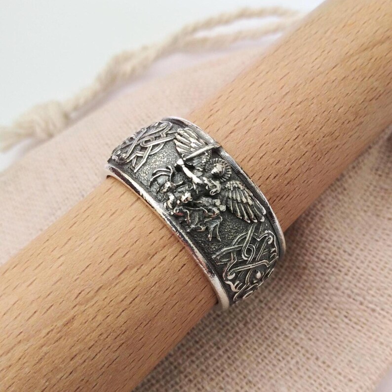 May include: A silver-colored ring with intricate embossed designs. The ring features a central motif of a winged creature and detailed Celtic knot patterns. The ring is shown on a wooden dowel.