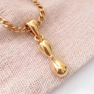Peut inclure: Un collier pendentif en métal doré composé d'une breloque lisse et polie en forme de cacahuète, suspendue à une chaîne à maillons gourmette assortie. Le pendentif présente une surface métallique réfléchissante au design arrondi et allongé.