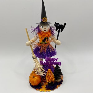 May include: A purple and orange Halloween witch doll with a black hat and broom. The doll is holding a black cat wand and is standing on a base with a pumpkin and pine trees. The base has the text "Happy Halloween" on it.