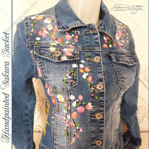 May include: A blue denim jacket with hand-painted pink, white, and green cherry blossoms. The jacket has a button closure and two pockets.