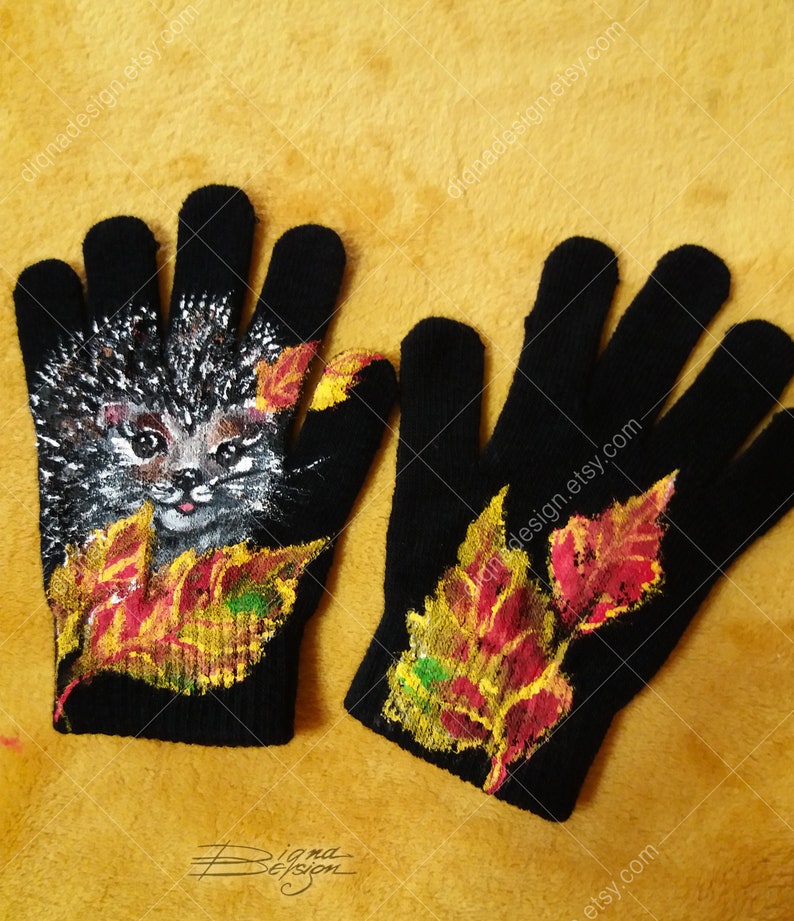 Puede incluir: Un par de guantes de punto negros con un dise&ntilde;o pintado a mano. Un guante presenta un erizo con hojas de oto&ntilde;o, mientras que el otro guante tiene un dise&ntilde;o de hojas de oto&ntilde;o. Los guantes est&aacute;n sobre un fondo amarillo.