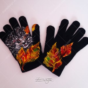Puede incluir: Un par de guantes de punto negros con un dise&ntilde;o pintado de un erizo y hojas de oto&ntilde;o. El erizo es gris y blanco con la nariz rosa y los ojos negros. Las hojas son rojas, naranjas, amarillas y verdes.