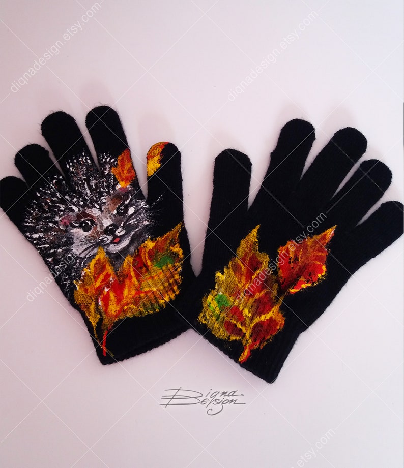 Puede incluir: Un par de guantes de punto negros con un dise&ntilde;o pintado de un erizo y hojas de oto&ntilde;o. El erizo es gris y blanco con la nariz rosa y los ojos negros. Las hojas son rojas, naranjas, amarillas y verdes.