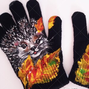 Puede incluir: Un par de guantes de punto negro con un dise&ntilde;o pintado de un erizo rodeado de hojas de oto&ntilde;o. El erizo tiene una cara linda y est&aacute; pintado en tonos de gris, marr&oacute;n y blanco. Las hojas est&aacute;n pintadas en tonos de naranja, amarillo y verde.
