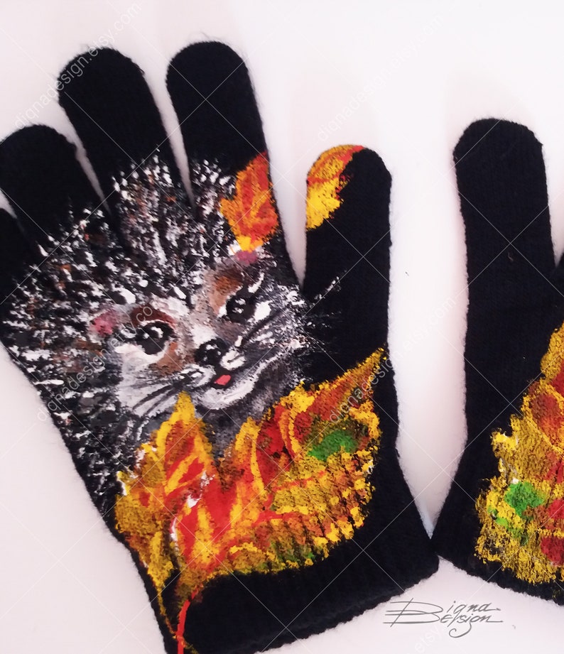 Puede incluir: Un par de guantes de punto negro con un dise&ntilde;o pintado de un erizo rodeado de hojas de oto&ntilde;o. El erizo tiene una cara linda y est&aacute; pintado en tonos de gris, marr&oacute;n y blanco. Las hojas est&aacute;n pintadas en tonos de naranja, amarillo y verde.