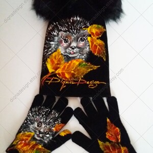 Puede incluir: Gorro negro de punto con pompones y guantes a juego. Tanto el gorro como los guantes presentan un dise&ntilde;o pintado de un erizo con hojas de oto&ntilde;o. El gorro tiene el texto "Digna Design" en la parte delantera.