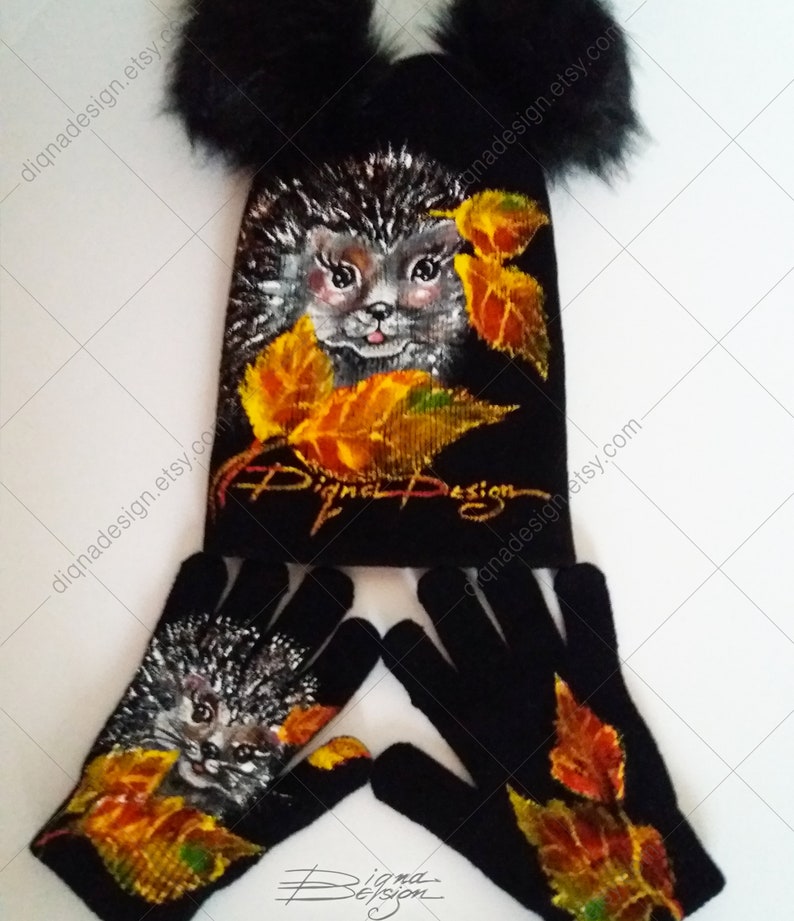 Puede incluir: Gorro negro de punto con pompones y guantes a juego. Tanto el gorro como los guantes presentan un dise&ntilde;o pintado de un erizo con hojas de oto&ntilde;o. El gorro tiene el texto "Digna Design" en la parte delantera.