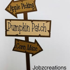 May include: A brown wooden signpost with three arrows pointing in different directions. The top arrow points left and says "Apple Picking". The middle arrow points right and says "Pumpkin Patch". The bottom arrow points right and says "Corn Maze". The signpost is on a white background with the text "Jobzcreations" below it.