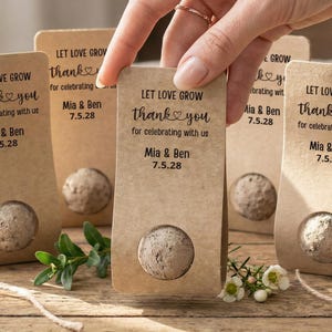 Pode incluir: Pacotes de sementes de papel kraft com o texto "Let Love Grow thank you for celebrating with us Mia & Ben 7.5.28". Cada pacote tem uma semente redonda. Lembrancinhas de casamento.
