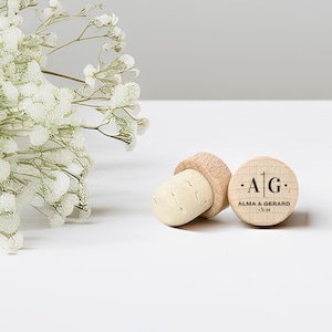 May include: Two wooden bottle stoppers with corks, one lying on its side and the other upright. The upright stopper has the initials "A | G" and the names "ALMA & GERARD" and a date printed on it. White flowers are in the background.
