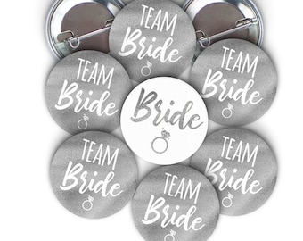 May include: A set of eight silver and white round buttons with the text "Team Bride" and "Bride" printed on them. The buttons are perfect for a bridal shower or bachelorette party.
