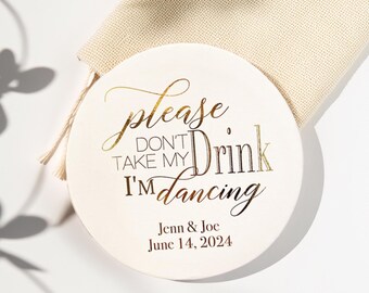 Könnte beinhalten: Ein runder, cremefarbener Getränkeuntersetzer mit goldfarbenem Schriftzug: "Please Don't Take My Drink I'm Dancing." Er trägt auch die Namen "Jenn & Joe" und das Datum "14. Juni 2024". Im Hintergrund ist ein beiger Beutel zu sehen.