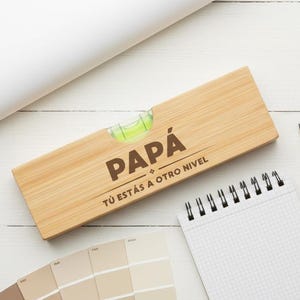 May include: A wooden spirit level with the word "PAPÁ" and the phrase "TÚ ESTÁS A OTRO NIVEL" printed on it, with a bubble level. A notepad, pencil, and paint swatches are on a white wooden surface.