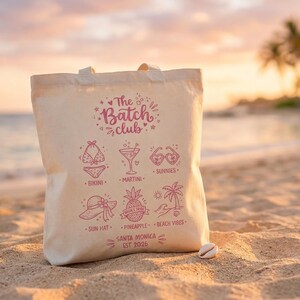 May include: A cream canvas tote bag with a pink design. It features beach-themed illustrations and the text "The Batch Club." The bag includes images of a bikini, martini, sunglasses, sun hat, pineapple, and palm trees. The text "Santa Monica EST 2026" is also included.