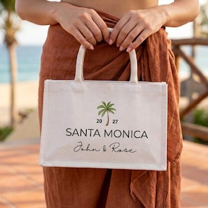 Könnte beinhalten: Eine weiße Canvas-Tragetasche mit cremefarbenen Griffen. Die Tasche zeigt eine grüne Palmen-Grafik und die schwarzen Worte "SANTA MONICA" sowie die Namen "John & Rose" in Schreibschrift. Die Zahlen "20 27" befinden sich über der Palme.