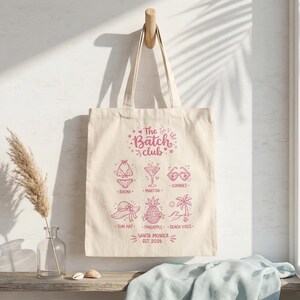 May include: Cream tote bag with a pink graphic design. The design includes beach-themed illustrations and text that reads "The Batch Club, Santa Monica, EST 2026". The bag is suspended from a wooden hook.