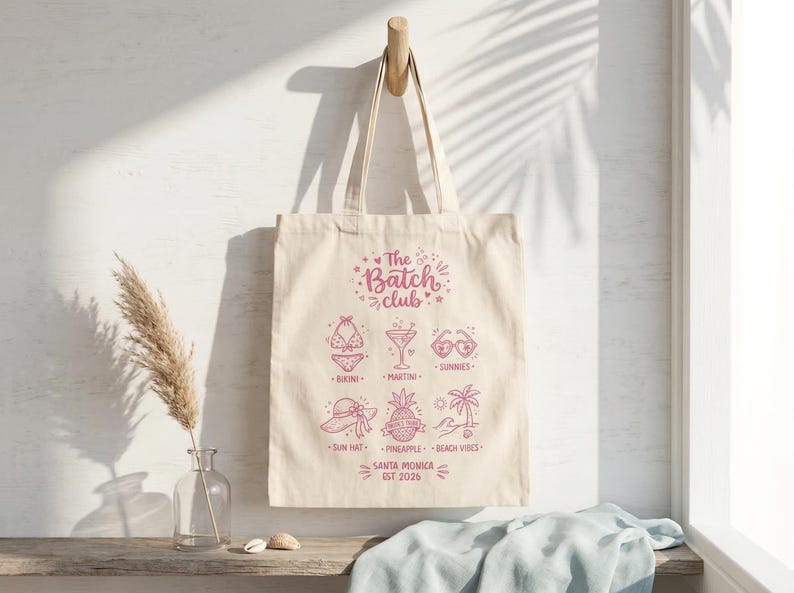 May include: Cream tote bag with a pink graphic design. The design includes beach-themed illustrations and text that reads "The Batch Club, Santa Monica, EST 2026". The bag is suspended from a wooden hook.