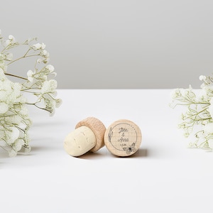 May include: Three wooden bottle stoppers. One stopper is engraved with the names "John & Anna", a butterfly, and the date 12.26. The stoppers are light brown and beige. The background is white with white flowers.