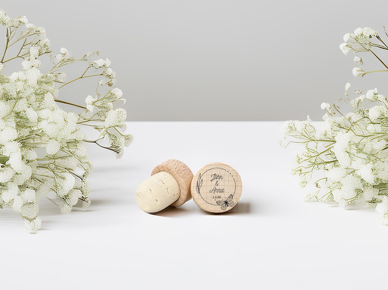 May include: Three wooden bottle stoppers. One stopper is engraved with the names "John & Anna", a butterfly, and the date 12.26. The stoppers are light brown and beige. The background is white with white flowers.