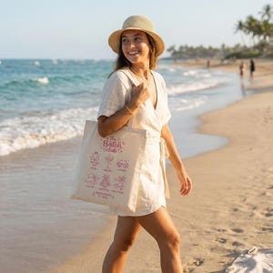 May include: A woman walks along a sandy beach, carrying a cream tote bag. The bag has pink cocktail illustrations and the text "The Batch Club". She wears a straw hat and a light-coloured wrap dress, enjoying the sunny day.