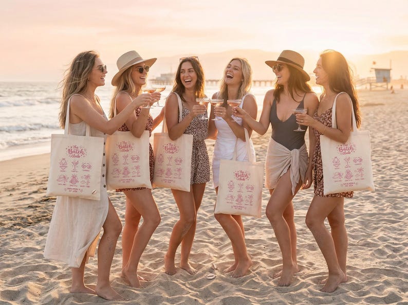 May include: A group of six people on a beach at sunset, raising glasses in a toast. Each person carries a canvas tote bag with "The Batch Club" and cocktail designs. The women are wearing dresses and swimwear.