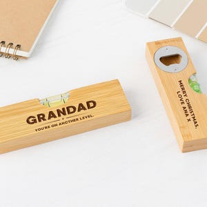 May include: Two wooden gifts on a white surface. One is a level with the text "GRANDAD" and "YOU'RE ON ANOTHER LEVEL". The other is a bottle opener with a silver opener and the text "MERRY CHRISTMAS, LOVE ANA X".