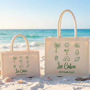 Könnte beinhalten: Zwei beigefarbene Tragetaschen mit grünen tropischen Illustrationen und dem Text "Los Cabos". Die Taschen befinden sich an einem Sandstrand mit dem Meer im Hintergrund. Die Taschen haben Griffe und sind mit einem Datum personalisiert.