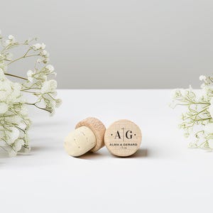 May include: Two wooden bottle stoppers with corks, one lying on its side and the other upright. The upright stopper has the initials "A G" and the names "Alma & Gerard" printed on it. White flowers are on either side.