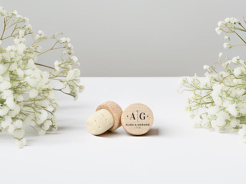 May include: Two wooden bottle stoppers with corks, one lying on its side and the other upright. The upright stopper has the initials "A G" and the names "Alma & Gerard" printed on it. White flowers are on either side.