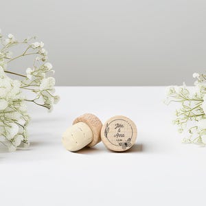 May include: Three wooden bottle stoppers with corks. One stopper is engraved with the names "John & Anna", a butterfly, and the date "12.26". The stoppers are on a white surface, with white flowers in the background.