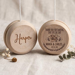 Può includere: Due yo-yo rotondi in legno appesi a cordini bianchi. Il yo-yo a sinistra è inciso con il nome Harper. Quello a destra presenta il testo Thank you for sharing our special day, i nomi María & Carlos, la data 07.12.28 e una coppia disegnata.