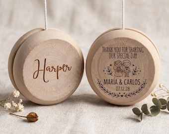 May include: Two round wooden yo-yos hanging by white strings. The left yo-yo is engraved with the name Harper. The right yo-yo features the text Thank you for sharing our special day, the names María & Carlos, the date 07.12.28, and a cartoon couple.