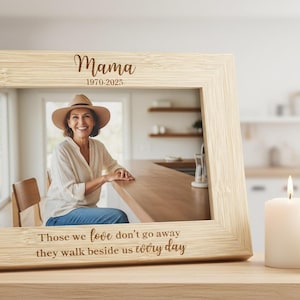 May include: A light-colored wooden picture frame with the word "Mama" and the years 1970-2025 engraved at the top. The frame holds a photo of a woman wearing a hat. The text "Those we love don't go away they walk beside us every day" is engraved at the bottom.