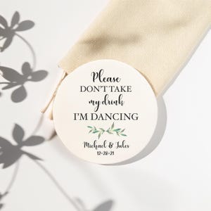 May include: A round, off-white coaster with the text "Please DON'T TAKE my drink I'M DANCING". Below the text are green leaves and the names "Michael & Jules" with the date "12-28-21". The coaster is next to a beige drawstring bag.