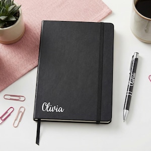 Könnte beinhalten: Ein schwarzes Notizbuch mit dem Namen "Olivia" in weißer Schrift, ein schwarzer Stift mit "Olivia" aufgedruckt und mehrere Büroklammern sind auf einer weißen Oberfläche angeordnet. Eine Tasse Kaffee und kleine Sukkulenten in Töpfen sind ebenfalls vorhanden.