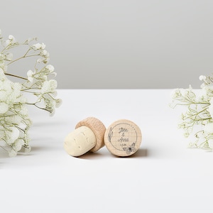 May include: Three wooden bottle stoppers with corks. One stopper is engraved with the names "John & Anna", a butterfly, and the date "12.26". The stoppers are on a white surface, with white flowers in the background.