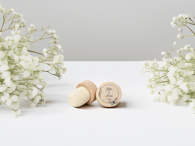 May include: Three wooden bottle stoppers with corks. One stopper is engraved with the names "John & Anna", a butterfly, and the date "12.26". The stoppers are on a white surface, with white flowers in the background.