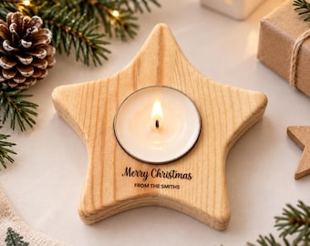 May include: A star-shaped light-coloured wooden tea light candle holder with a natural wood grain finish. The centre holds a lit white tea light candle. The text Merry Christmas FROM THE SMITHS is engraved in black on the front of the wooden star.