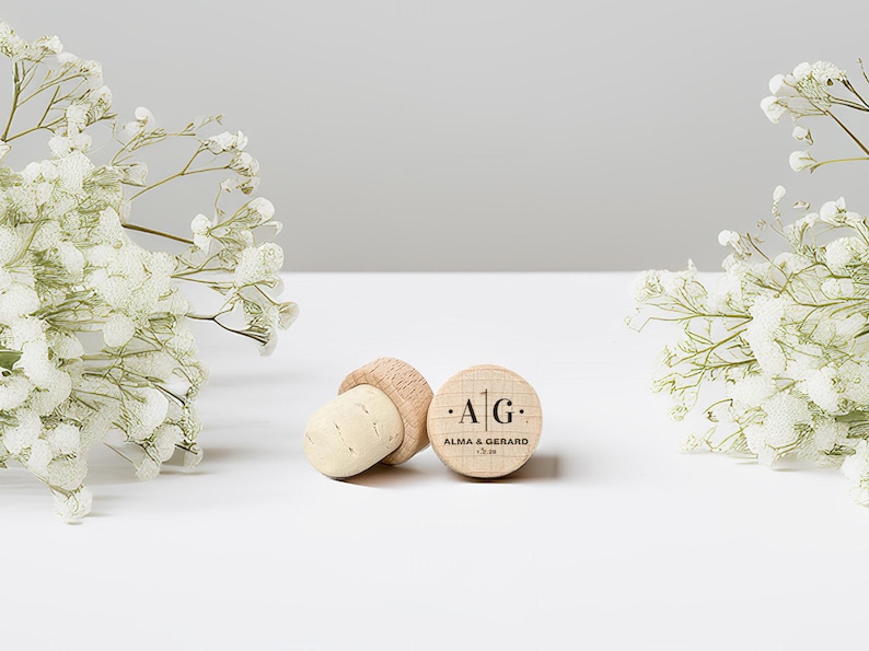 May include: Two wooden bottle stoppers with corks, one lying on its side and the other upright. The upright stopper has the initials "A | G" and the names "ALMA & GERARD" and a date printed on it. White flowers are in the background.