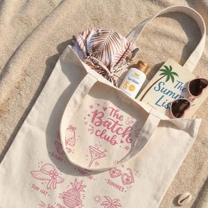May include: A cream tote bag containing summer items. It holds a bikini, sunscreen, a book titled "The Summer List," and heart-shaped sunglasses. The bag has pink illustrations and the text "The Batch Club."
