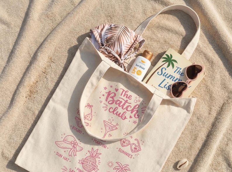 May include: A cream tote bag containing summer items. It holds a bikini, sunscreen, a book titled "The Summer List," and heart-shaped sunglasses. The bag has pink illustrations and the text "The Batch Club."