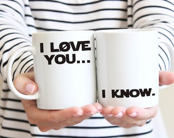 Może przedstawiać: Dwa białe kubki do kawy z czarnym tekstem. Kubek po lewej stronie ma napis "I LOVE YOU...", a kubek po prawej stronie ma napis "I KNOW."