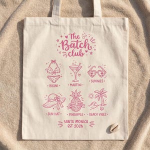 Könnte beinhalten: Eine cremefarbene Tragetasche mit einem rosa Strandmotiv. Das Design umfasst Illustrationen eines Bikinis, eines Martinis, einer Sonnenbrille, eines Sonnenhuts, einer Ananas und Palmen. Der Text lautet "The Batch Club" und "Santa Monica EST 2026".