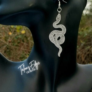 May include: Silver snake dangle earrings. The snake is coiled and has a detailed, textured design. The earrings are on a black mannequin.
