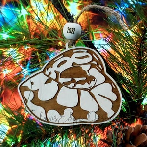 May include: A wooden Christmas ornament shaped like a snake with a white and brown pattern. The ornament is hanging on a green pine tree branch with a 2022 bead on the string.