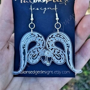 May include: Clear acrylic earrings featuring a pair of intertwined snakes. The earrings are silver and have a hook closure. The earrings are on a black card with the text "Talonsedge Designs" and the website address "talonsedgedesigns.etsy.com".