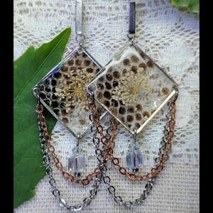 May include: A pair of handmade earrings with a clear, diamond-shaped resin design. The earrings feature dried flowers and a honeycomb pattern. Silver-tone metal accents and chains in silver and copper tones dangle from the bottom, with a clear crystal bead.